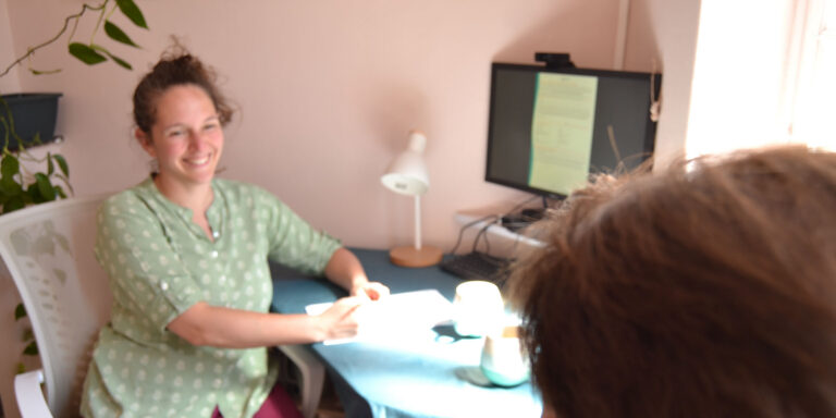 Consultation ayurvédique en cabinet à Saint-Girons. Praticienne a son bureau, elle sourit à un client lors d'une consultation ayurvédique de suivi. Consult an ayurvedic practitioner in person.