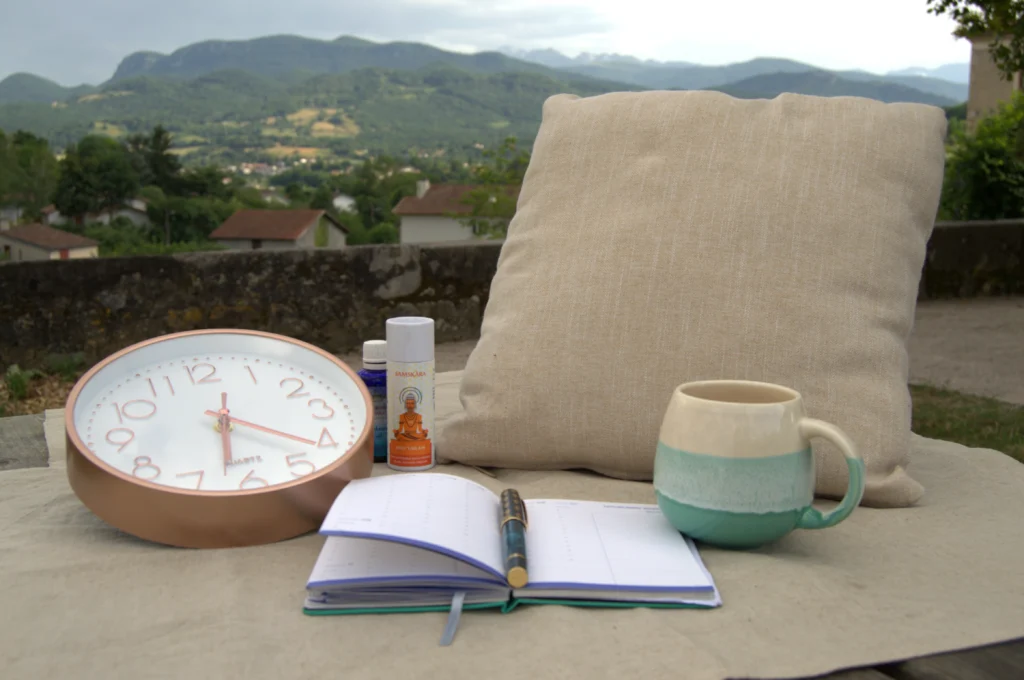 Une horloge, un coussin, un agenda, une tasse, des huiles hydratantes. Le paysage du couserans au fond
