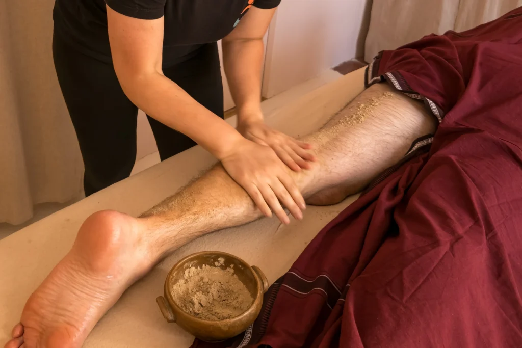 La praticienne frotte vigoureusement une poudre de plantes sur la jambe d'un client (soin Udvartana stimulant et drainant)